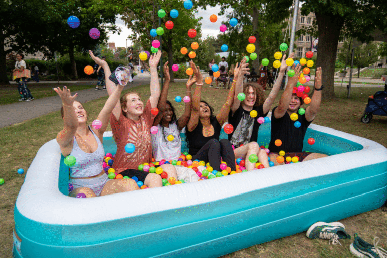 Group of students sitting in inflatable pool of colourful balls, throwing them in the air.