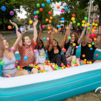 Group of students sitting in inflatable pool of colourful balls, throwing them in the air.